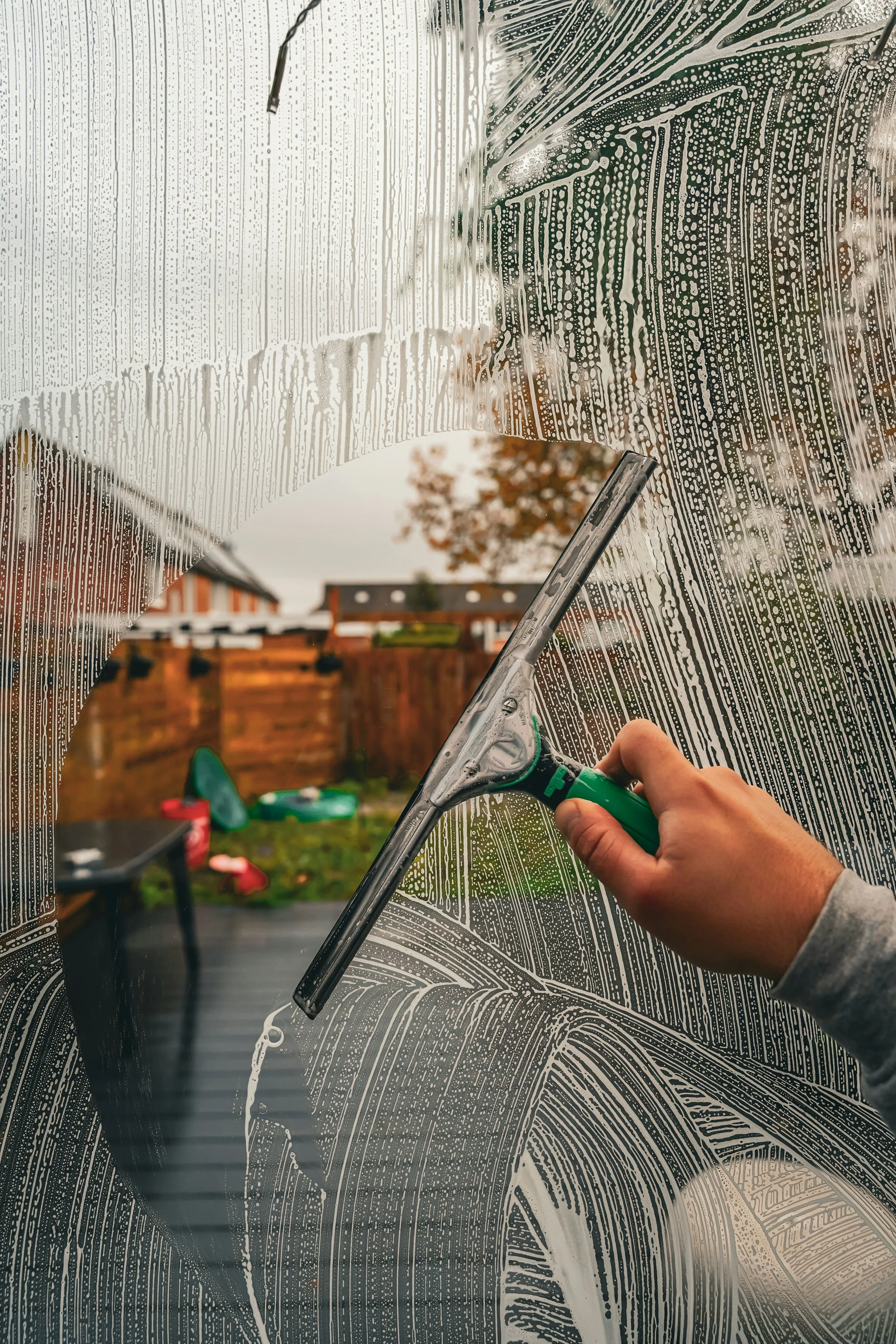 Person putzt eine wetterfeste PVC-Folie mit einem Scheibenwischer auf einer Glasscheibe. Flüssigkeit und Seifenreste sind auf der Scheibe sichtbar.
