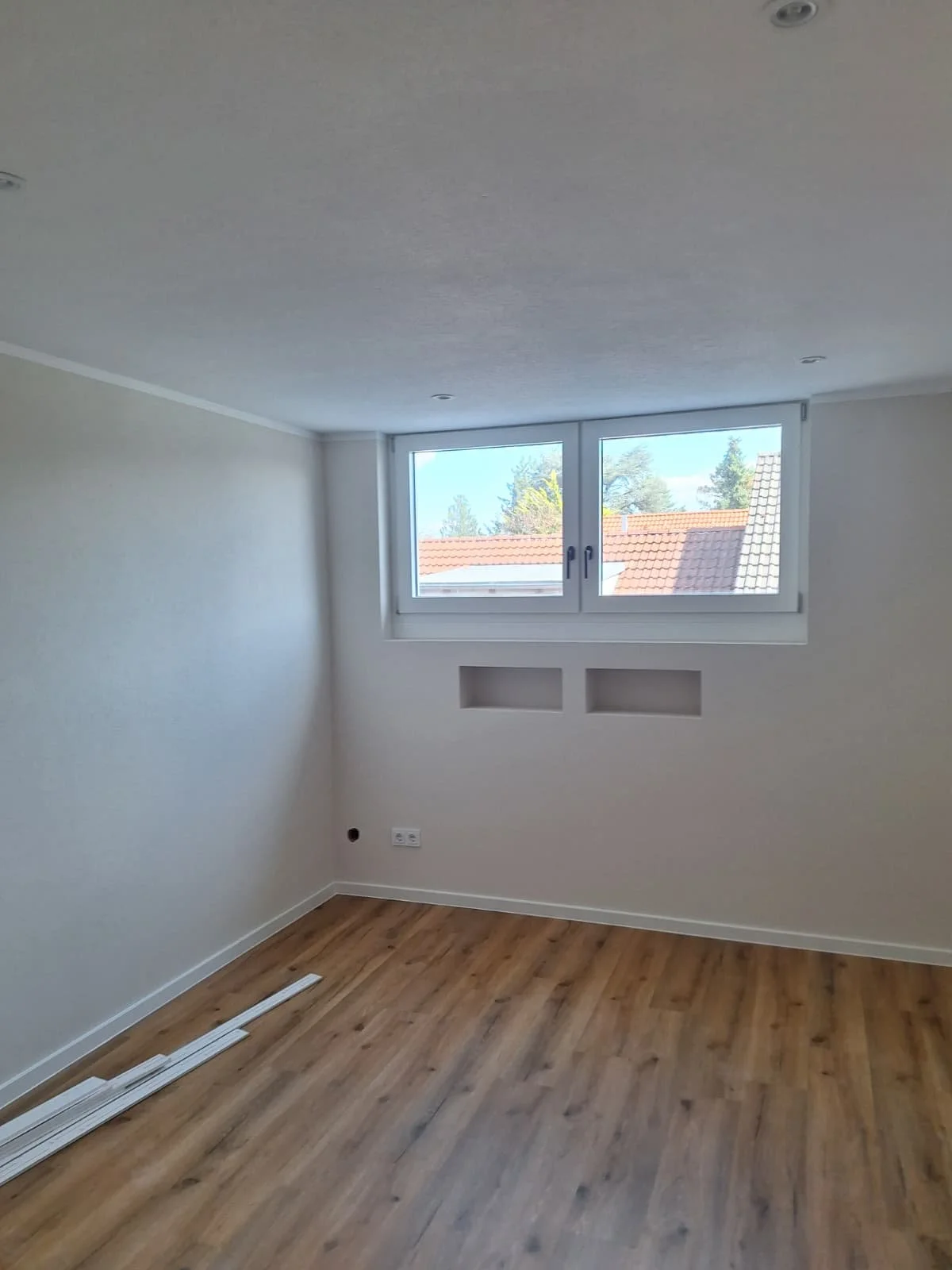 Ein leeres, renoviertes Zimmer mit weißen Wänden, Holzboden, einer großen Fensterfront und kleinen Wandnischen.