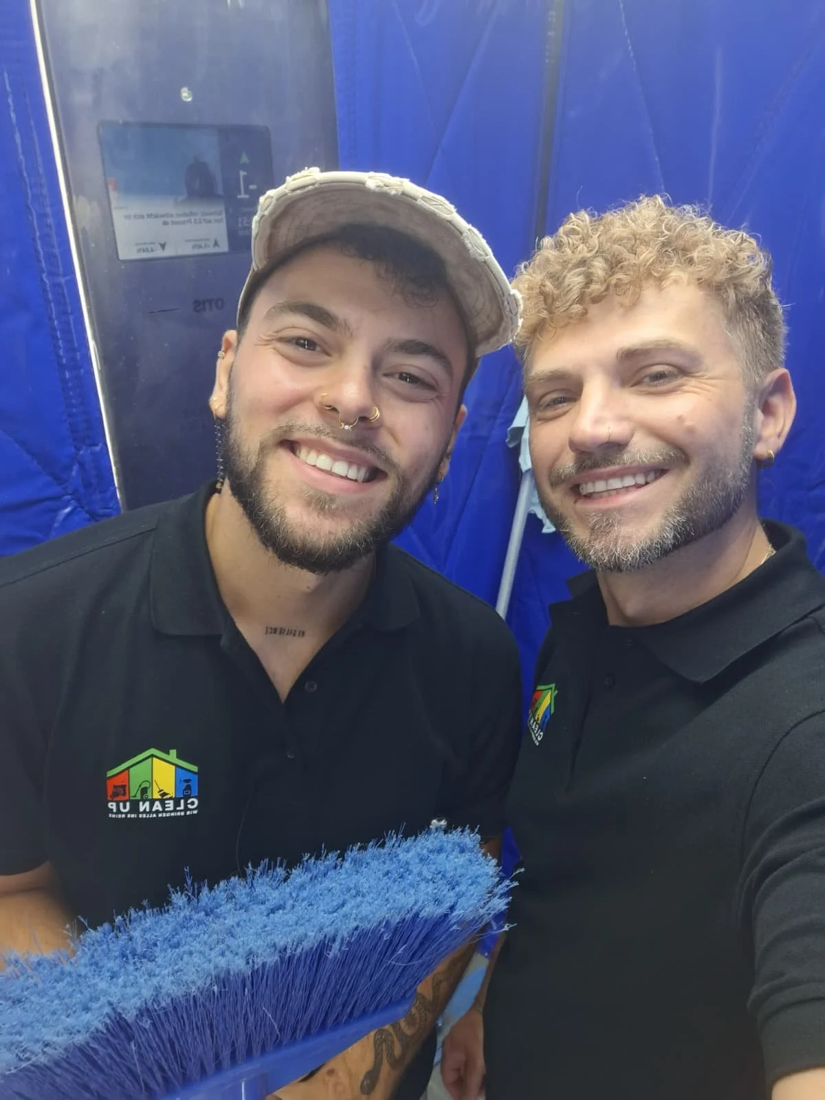 Zwei lachende Männer, die für ein Selfie posieren. Beide tragen schwarze T-Shirts mit einem bunten Logo, das ein Haus mit vier Farben zeigt. Sie sind in einem blauen Zelt oder Container, einer hält einen blauen Reinigungsbesen. Der Mann auf der linke