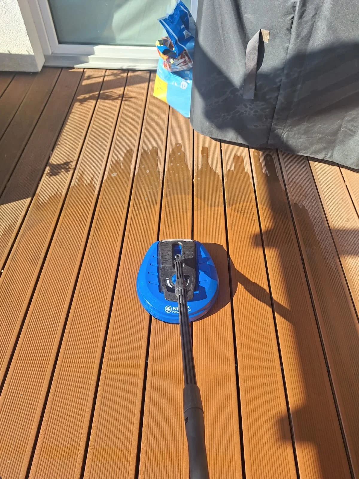 Der Blick auf einen Holz-Terrassenboden, während jemand mit einem blauen und schwarzen Reinigungsgerät eine Reinigung durchführt. Im Hintergrund sind eine geschlossene Tür und eine Tasche sichtbar.
