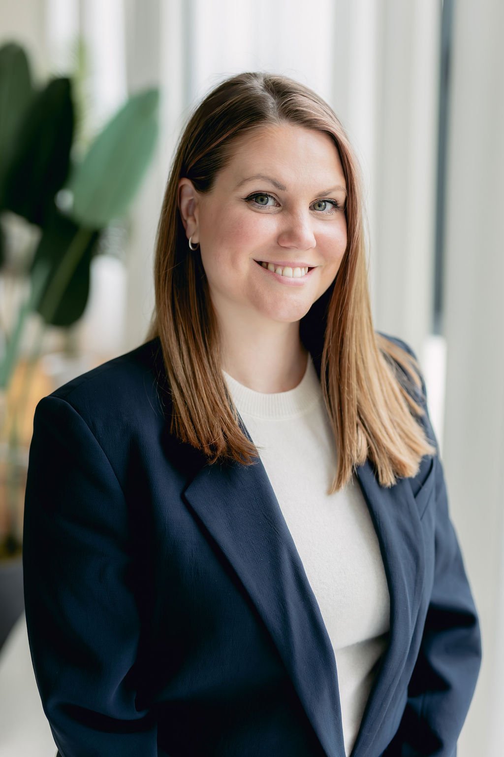 Katie Baker, headshot. 
A smiling woman with shoulder-length brown hair, wearing a navy blazer and a white top, standing in a bright office with large windows and green plants.