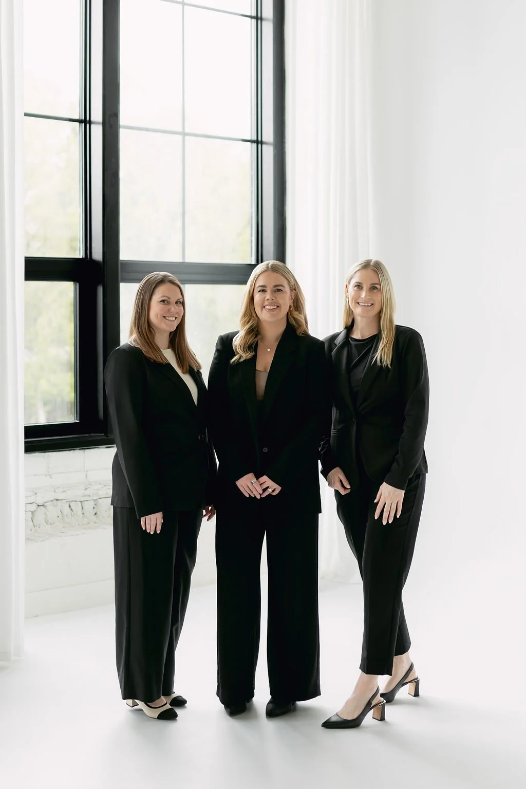 Katie Baker, Laura Smith and Kaitlin Jagersky.
Three women in black business suits standing in a bright, modern office with large window panels and white curtains.