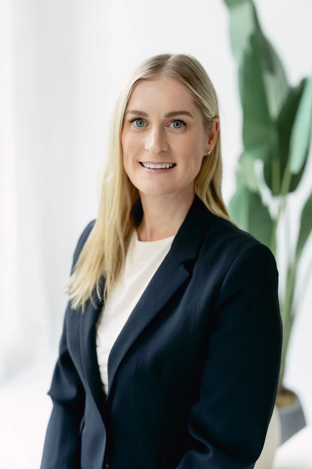 Kaitlin Jagersky, Headshot. A professional woman with blonde hair and blue eyes, wearing a black blazer and white shirt, smiling at the camera in an office setting with a plant in the background.