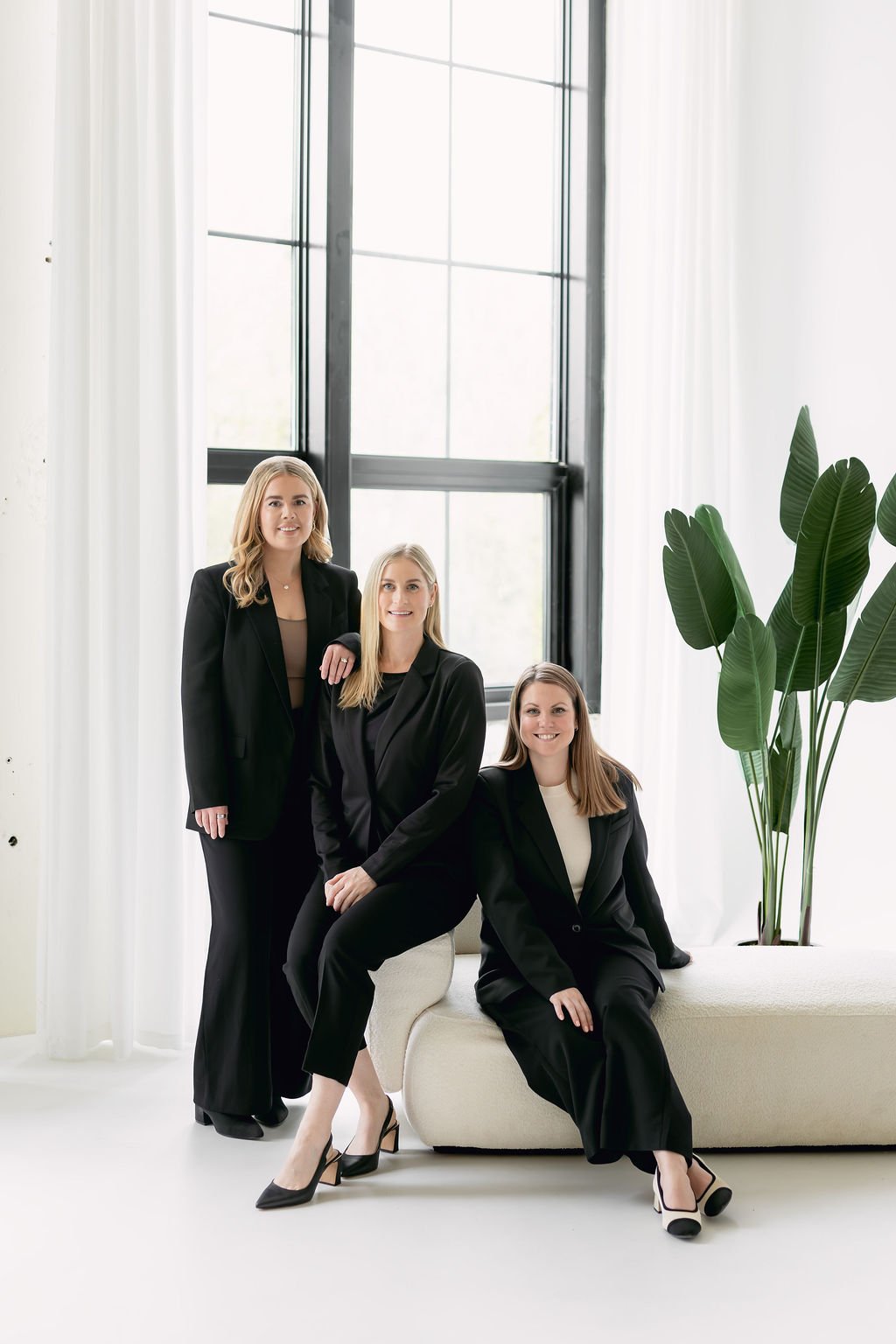 Katie Baker, Kaitlin Jagersky and Laura Smith. Three women in black business suits are in a modern office space with large windows, white curtains, and a tall green plant, posing together.