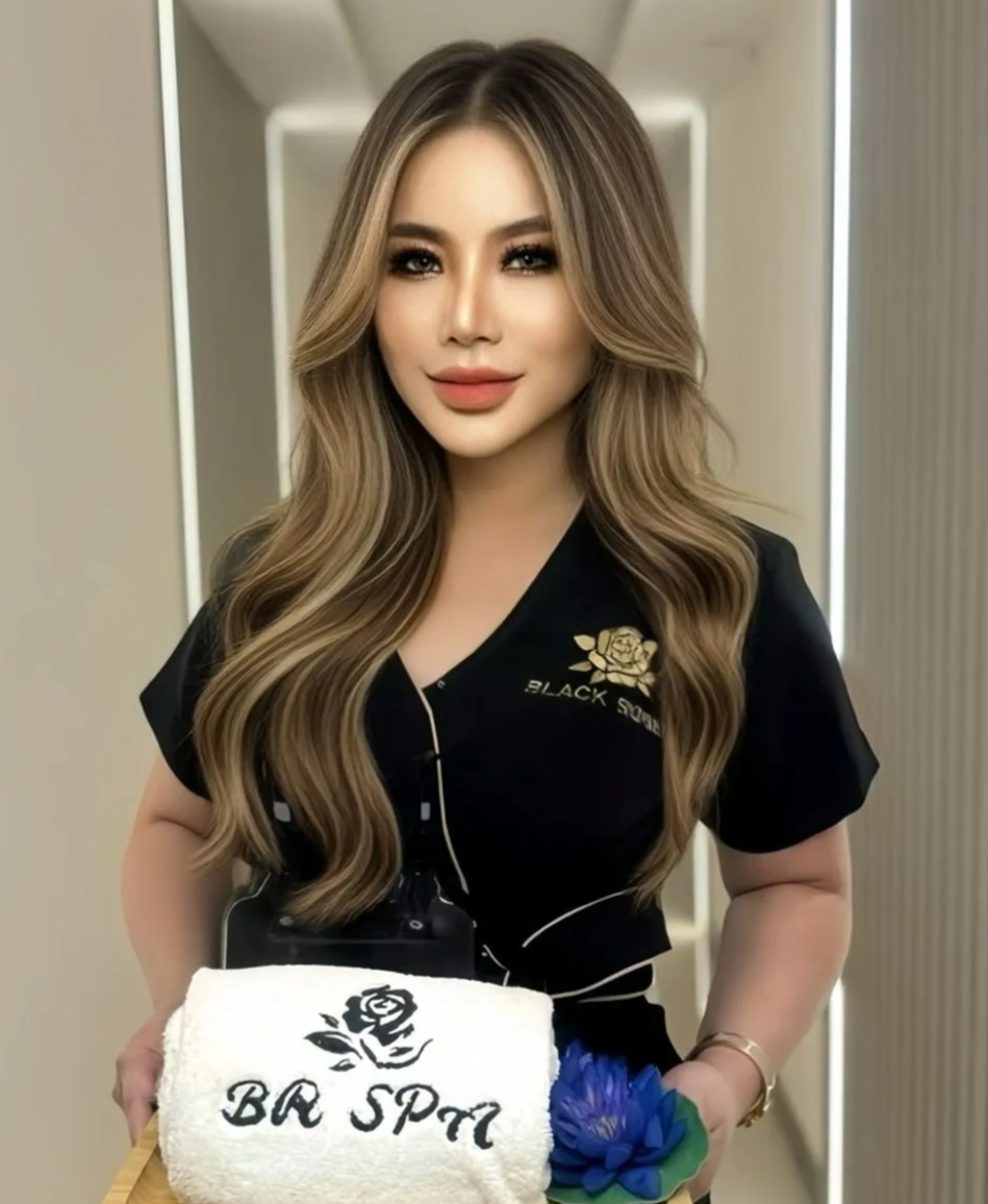 A woman with long, wavy blonde hair and makeup, wearing a black uniform with a gold embroidered rose and the words 'Black Spa' embroidered on it, holding a rolled white towel with a black rose and the words 'BRA SPA' written on it, and a blue flower. She is standing in a well-lit hallway.