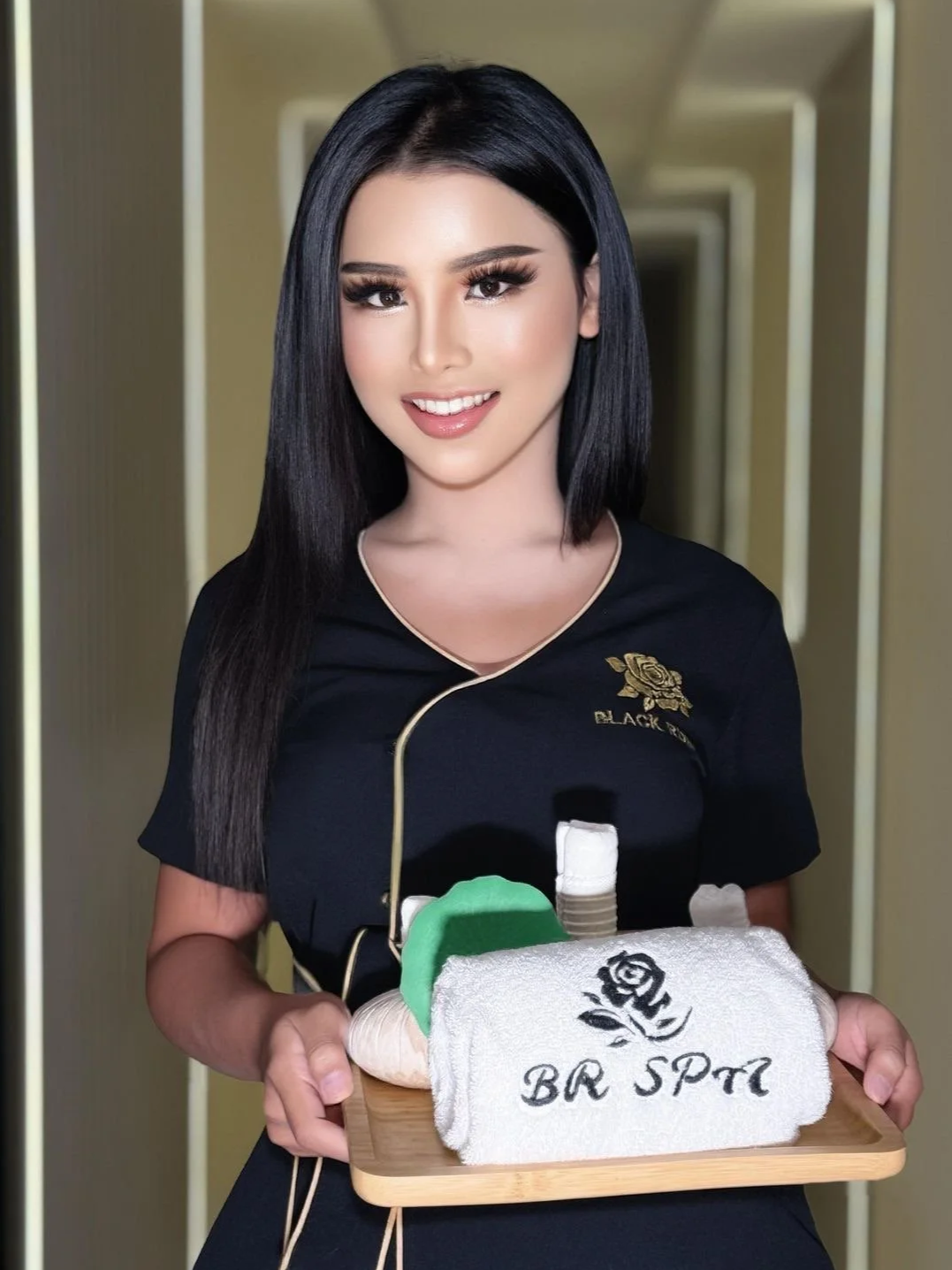 A woman with long black hair, dressed in black, holds a tray with a rolled towel embroidered with 'BR SPA,' a small bottle, and a green object, in a hallway.