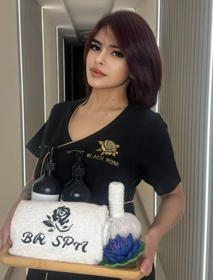 A woman in a black uniform with a gold rose logo and the words "Black Rose" stands in a hallway holding a tray with spa products and a towel. The tray contains a rolled white towel, two black bottles, a decorative soap wrapping, and blue and green decorative flowers.