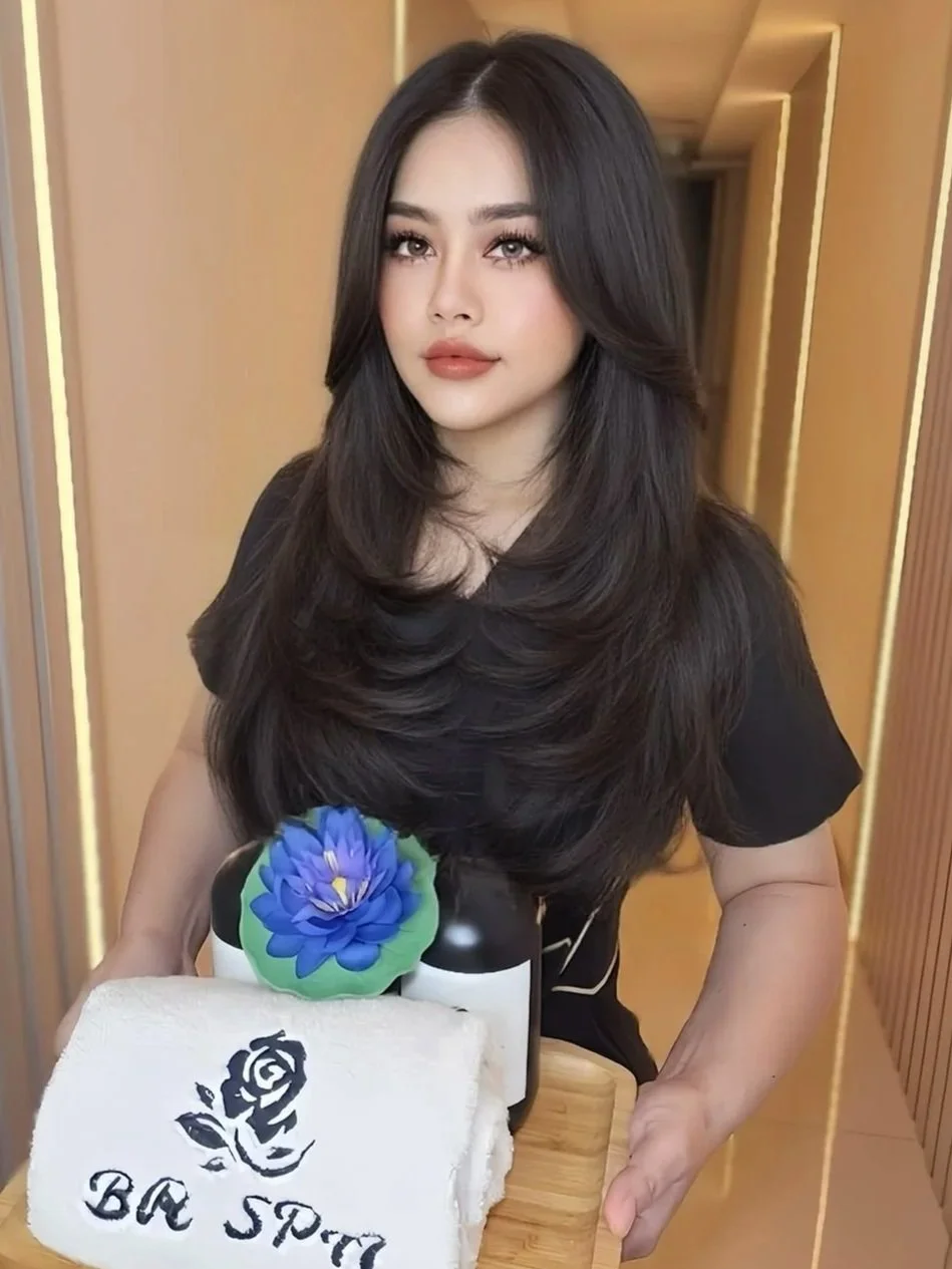A woman with long, dark hair and makeup stands in a hallway, holding a birthday cake with a white towel, a blue flower, and skincare products on it.