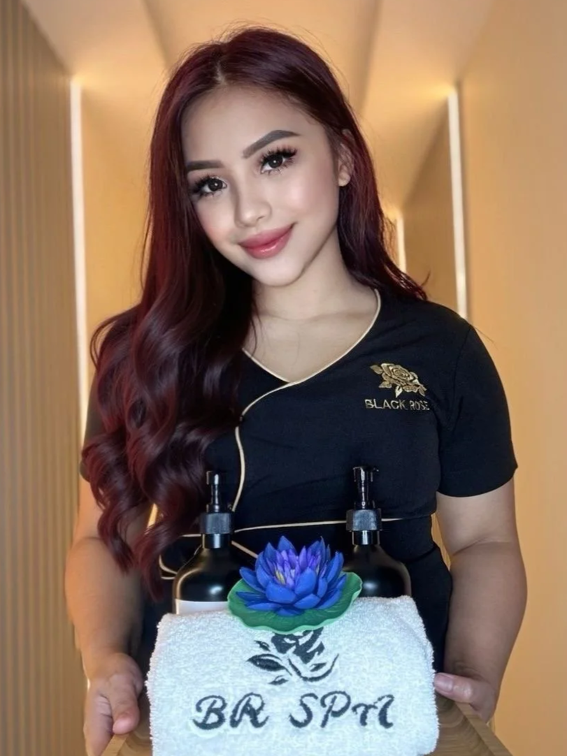 A woman with long wavy burgundy hair, wearing a black top with gold detailing and a logo that reads 'Black Rose,' stands indoors in front of warm lighting. She is holding two black pumps with soap dispensers on top, attached to a white towel with a blue lotus flower and the words 'BB Spa.'