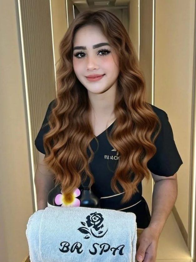 A young woman with long, wavy, light brown hair and makeup, smiling while holding a towel with a rose and the words 'BR SPA' on it, standing in a spa or salon corridor.