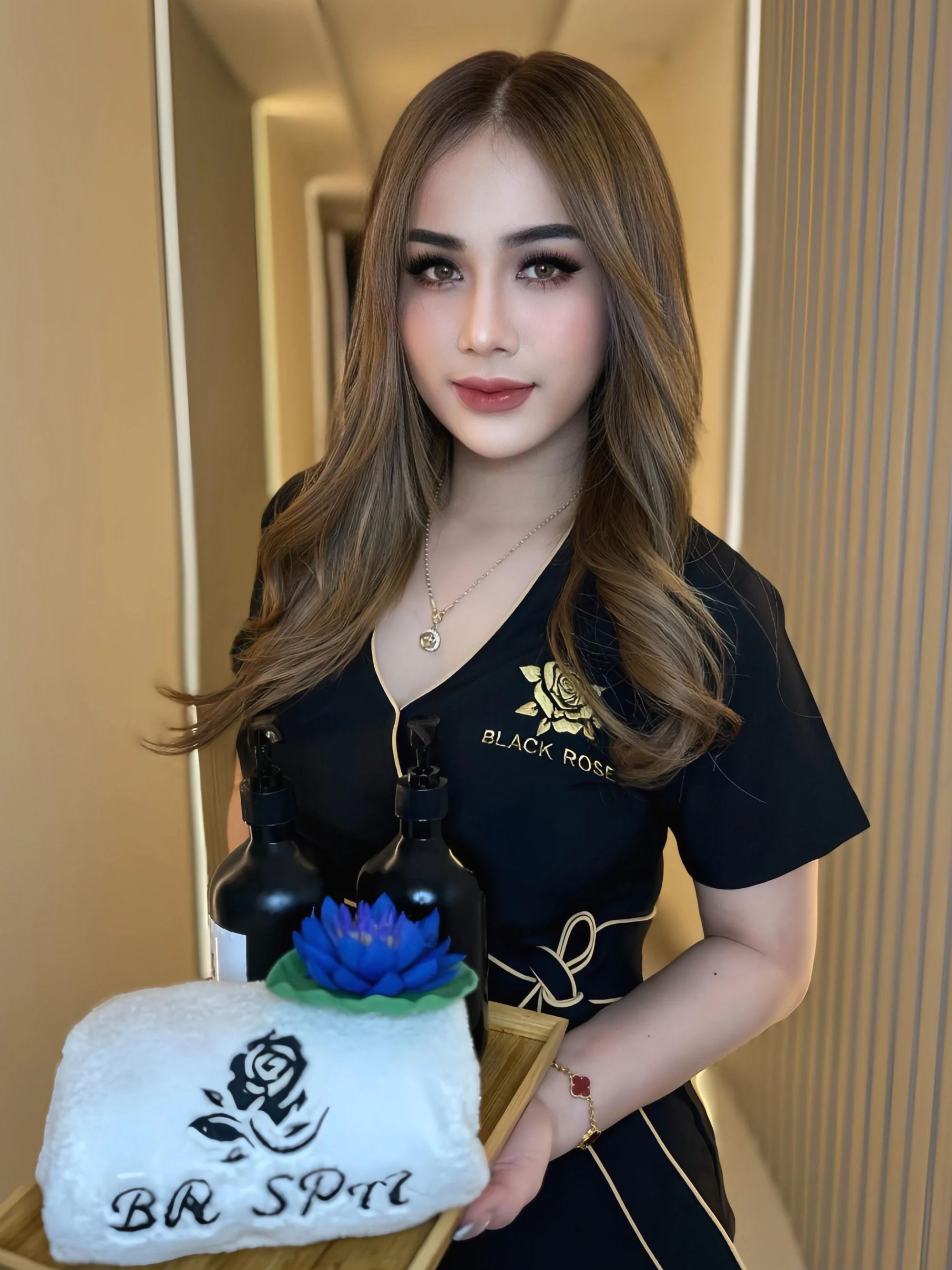 A woman with long wavy hair, wearing makeup and a black top with gold trim, stands behind a tray holding a towel with a rose and the text 'BB Spa'. The towel covers two black bottles with blue flowers on top. The background is a softly lit room with beige and gold accents.