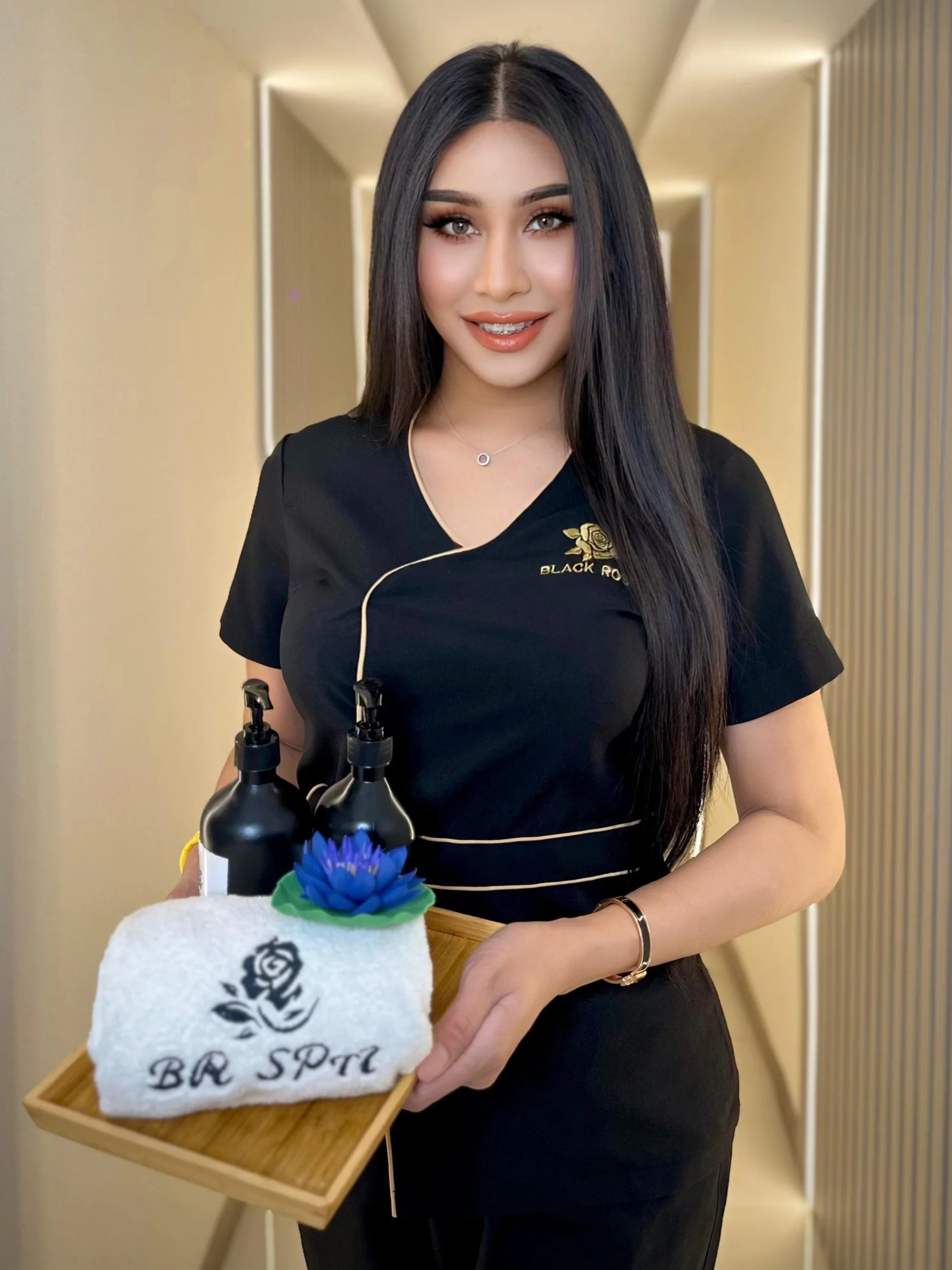 A woman with long black hair and light makeup, wearing a black uniform with gold accents, standing in a modern room, holding a wooden tray with a white towel embroidered with a black rose and the words 'BB SPA', and two black bottles with a blue flower decoration.