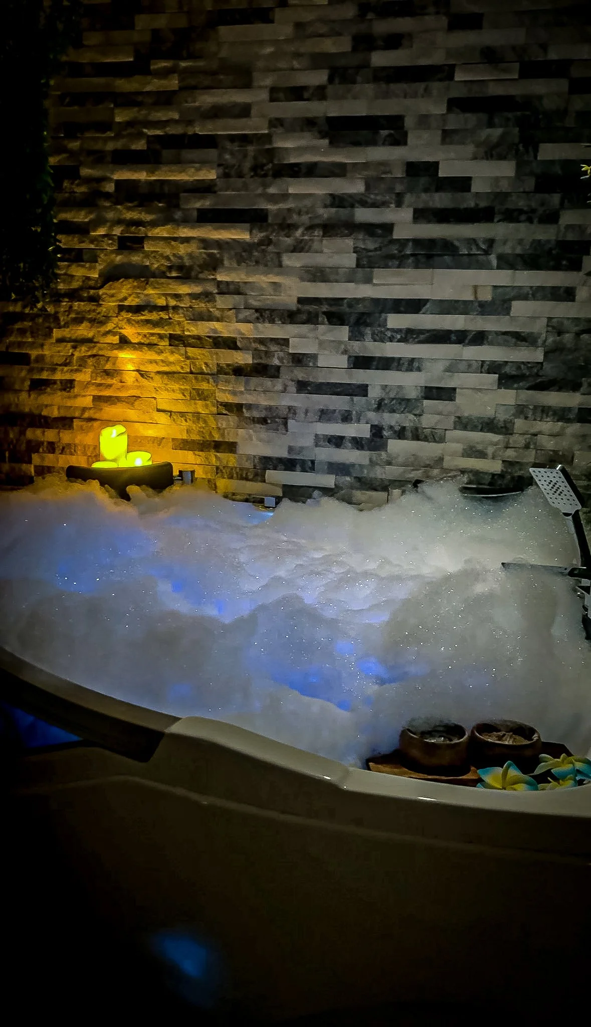 Night view of a bathtub filled with water, snow on the edges, and a stone wall with warm lighting in the background, with slippers and flowers on the edge.