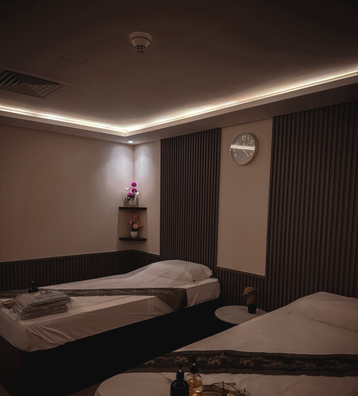 Modern couple spa room with two single beds, bedside table, wall clock, decorative lighting, and floral decor.