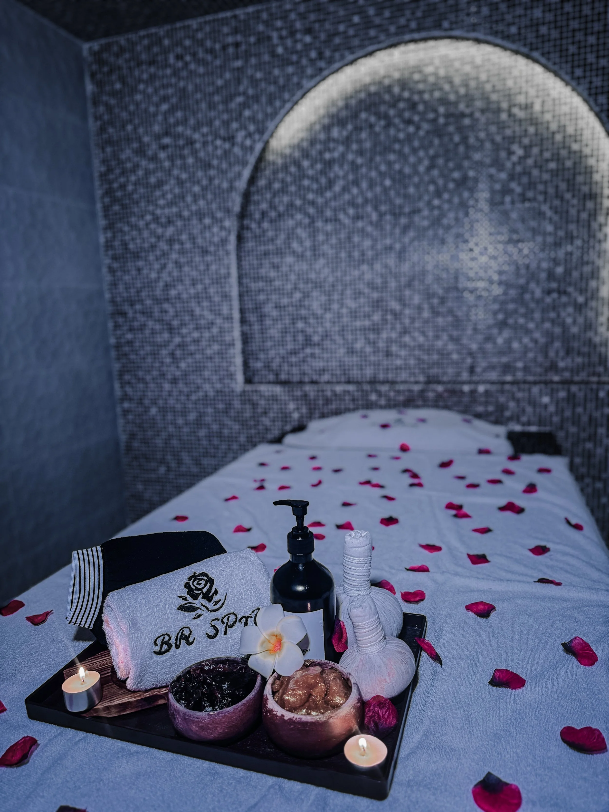 A romantic spa setting with rose petals scattered on a bed, a tray with candles, spa products, and flowers, and folded towels with spa branding.