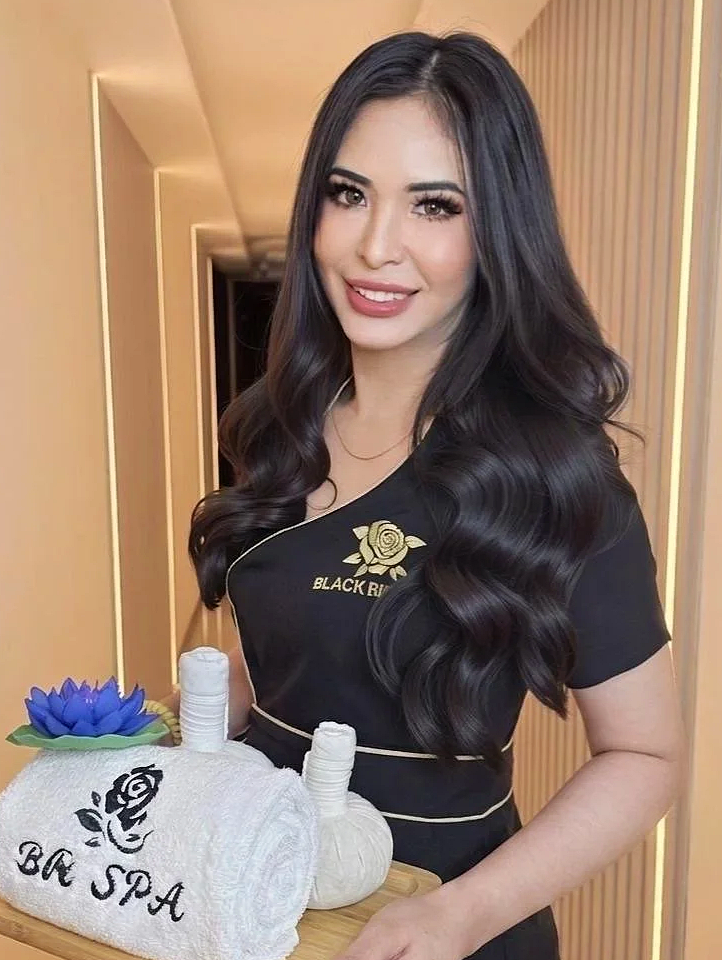 Young woman with long, wavy black hair smiling while standing in a spa, holding a tray with towels, a small sculpture, and a flower, wearing a black uniform with gold embroidery that reads 'Black Rose'.