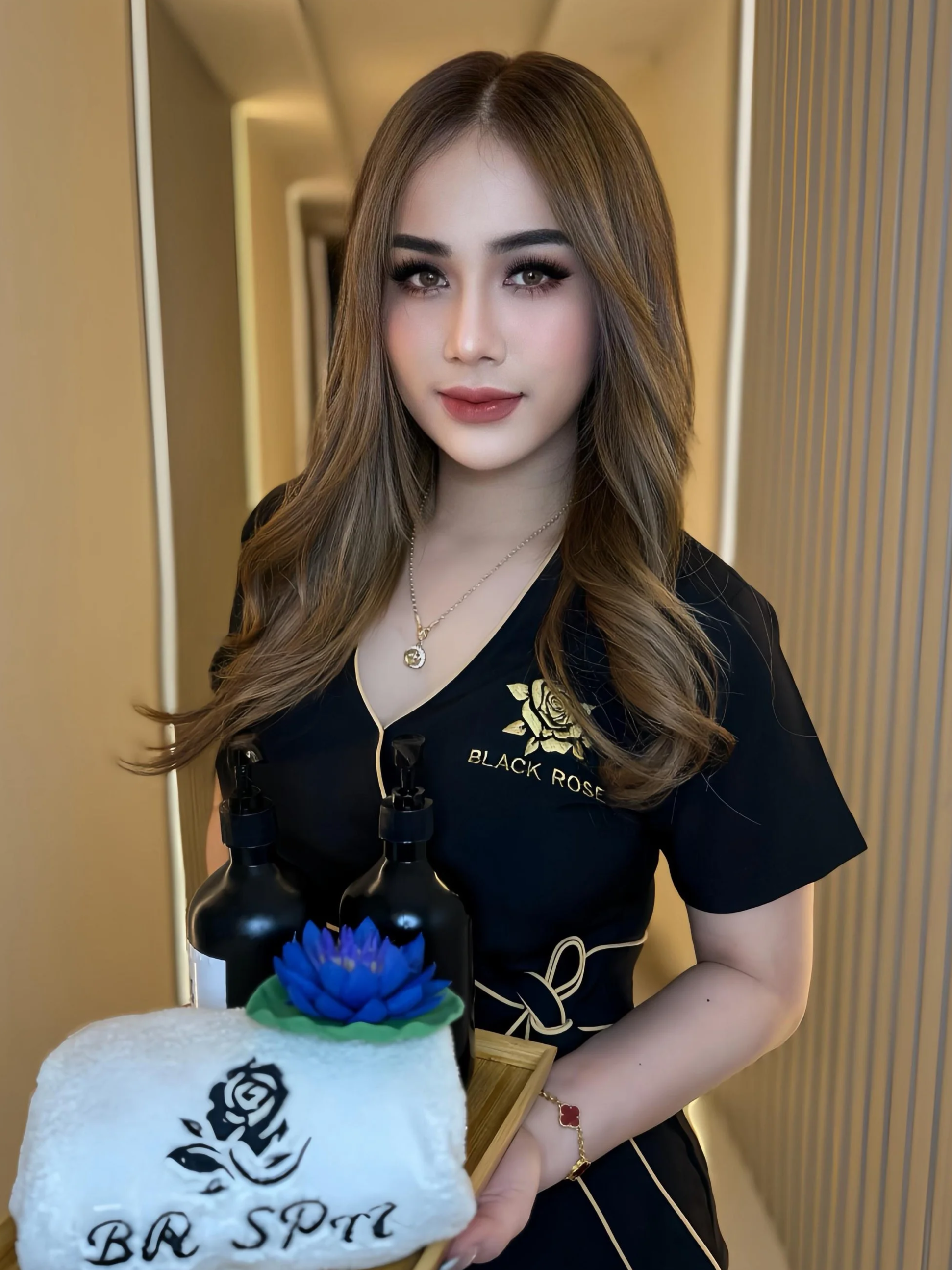 A young woman with long, wavy light brown hair and makeup, wearing a black shirt with gold embroidery, holding a tray with bottles and a white towel with a black rose design and the words BR Spa, standing in a hallway with beige walls and wooden accents.