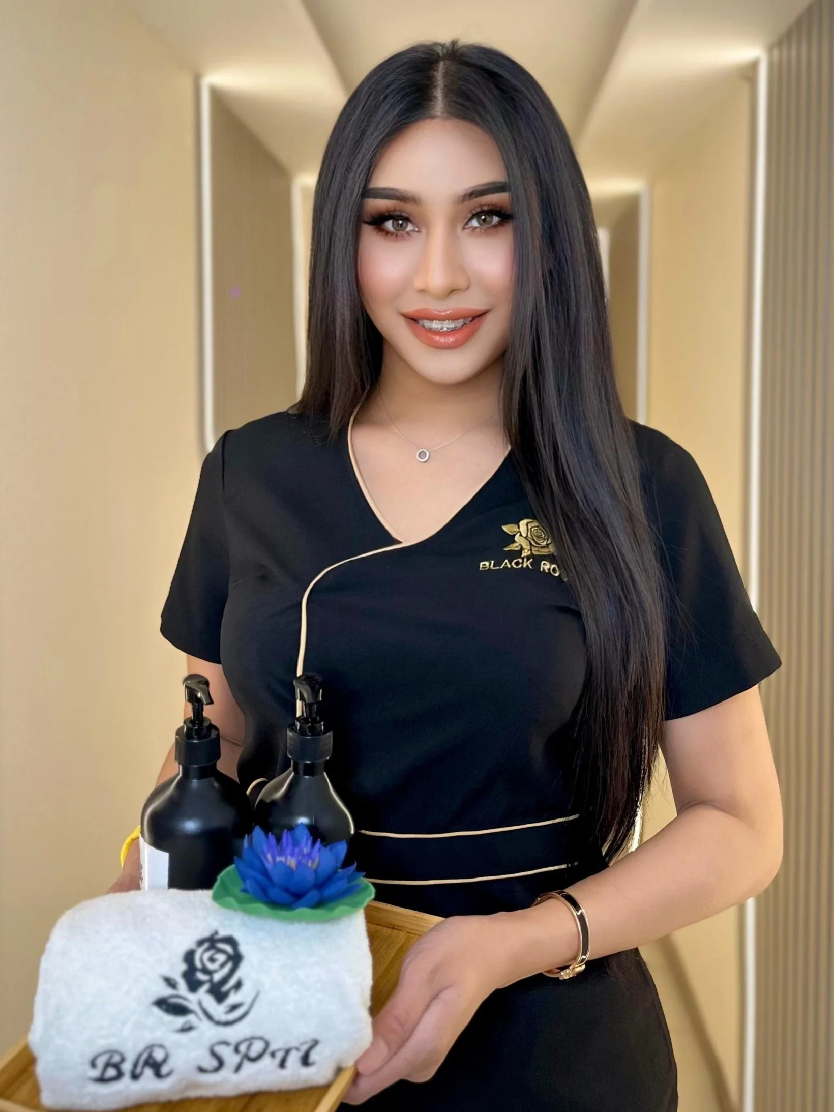 A woman with long dark hair and makeup, wearing a black shirt with gold embroidery, holding a towel with a logo, a wooden tray with bottles, a blue flower, and a spa ring, in a spa or wellness setting.
