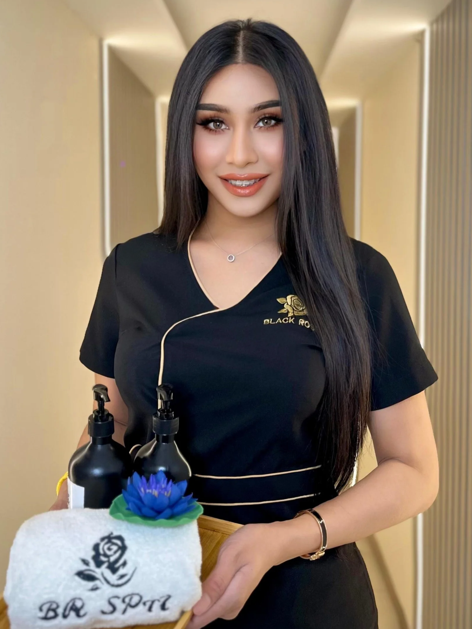 A smiling woman with long black hair and makeup, wearing a black uniform with 'Black Rose' embroidered on it, holding a towel with a black rose logo and 'BR SP TL' text, and a small decorative blue flower, with a spa or wellness center background.