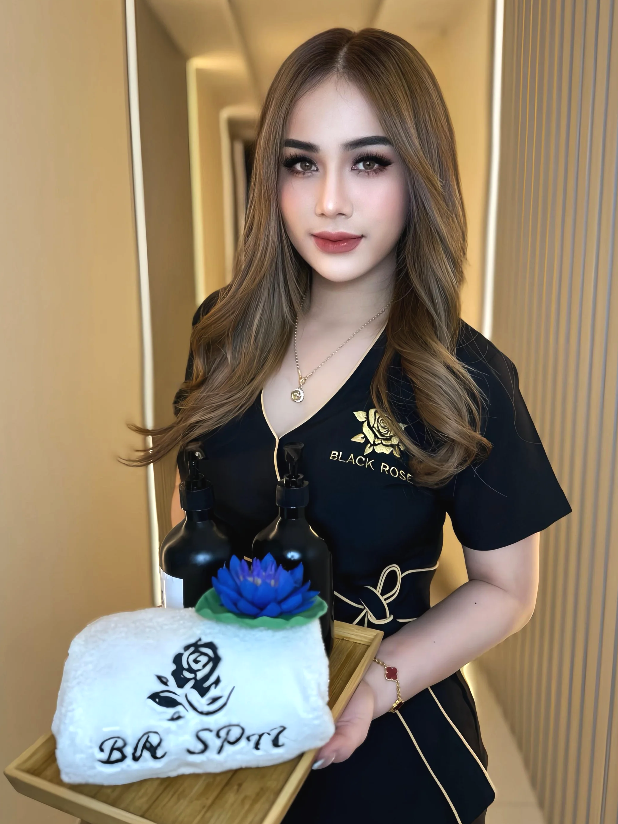 A woman with long, wavy hair and makeup standing in an indoor setting, wearing a black shirt with a gold logo that reads 'Black Rose,' holding a wooden tray with a white stone engraved with a black rose and the words 'BE SPOT' and two black bottles. The background features a warm-colored wall and lighting.