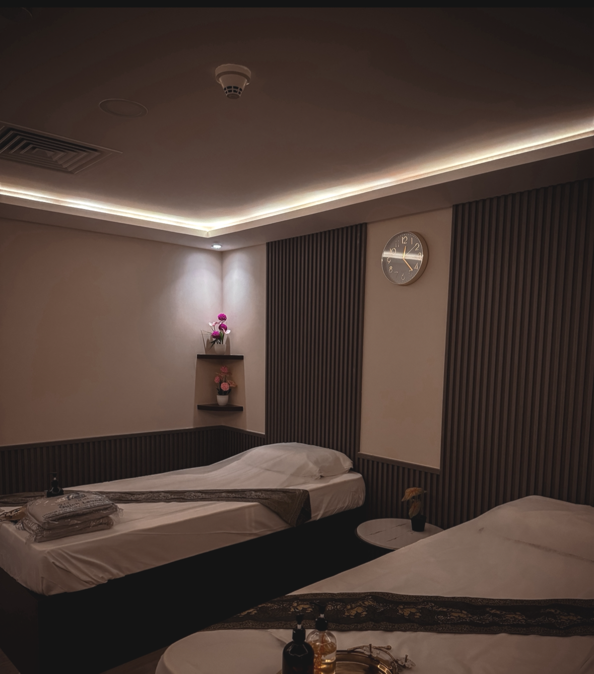 Modern couple spa room with two single beds, bedside table, wall clock, decorative lighting, and floral decor.