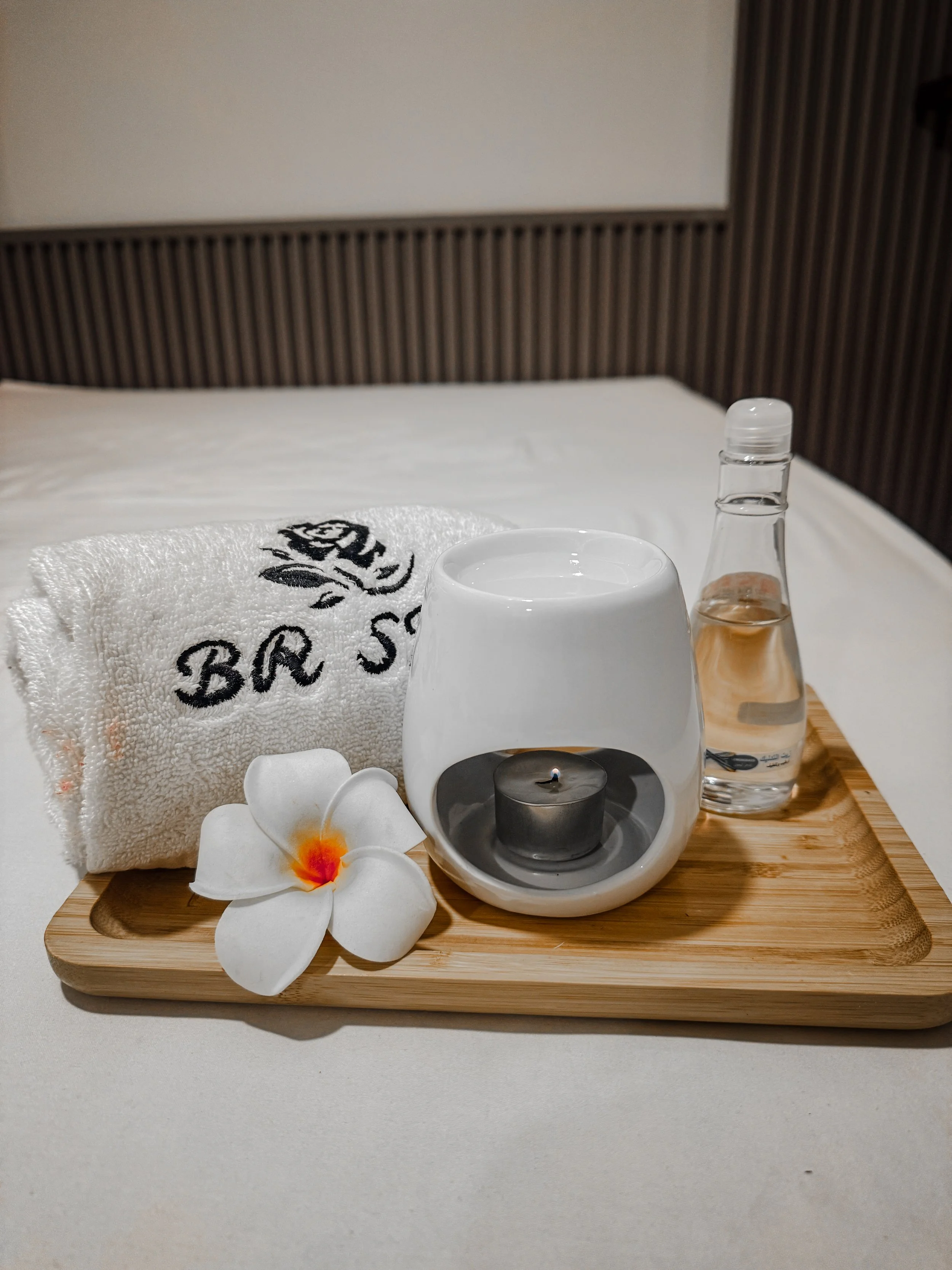 A wooden tray with a white towel embroidered with black text, a small white candle holder with a tealight candle, a glass bottle of lotion or oil, a white ceramic bowl, a white flower with orange center, and some small white stones. The tray is on a white surface with a bed and headboard in the background.
