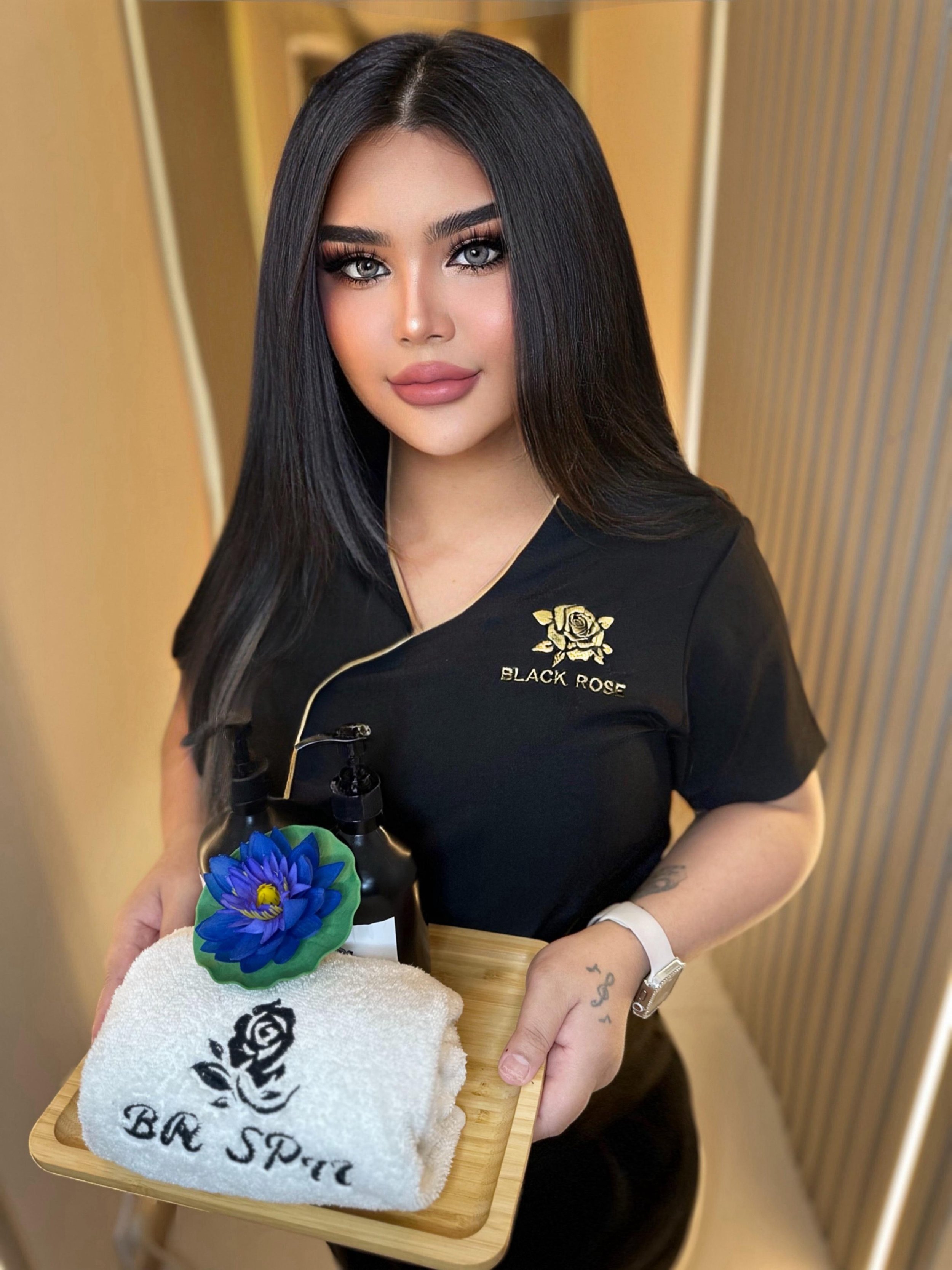 A woman with long black hair, gray eyes, and makeup, wearing a black shirt with a gold rose and the words "Black Rose" embroidered on it, stands behind a wooden table. She is holding a towel with a black rose logo and the words "Be Spa" embroidered, and a blue flower decoration. The background features a wood-paneled wall.