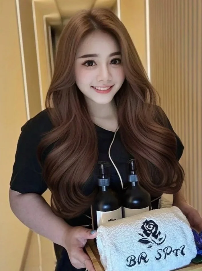 Young woman with long wavy brown hair smiling, holding a tray with two bottles, a rolled towel with a rose and the words 'BR SPa' embroidered on it.