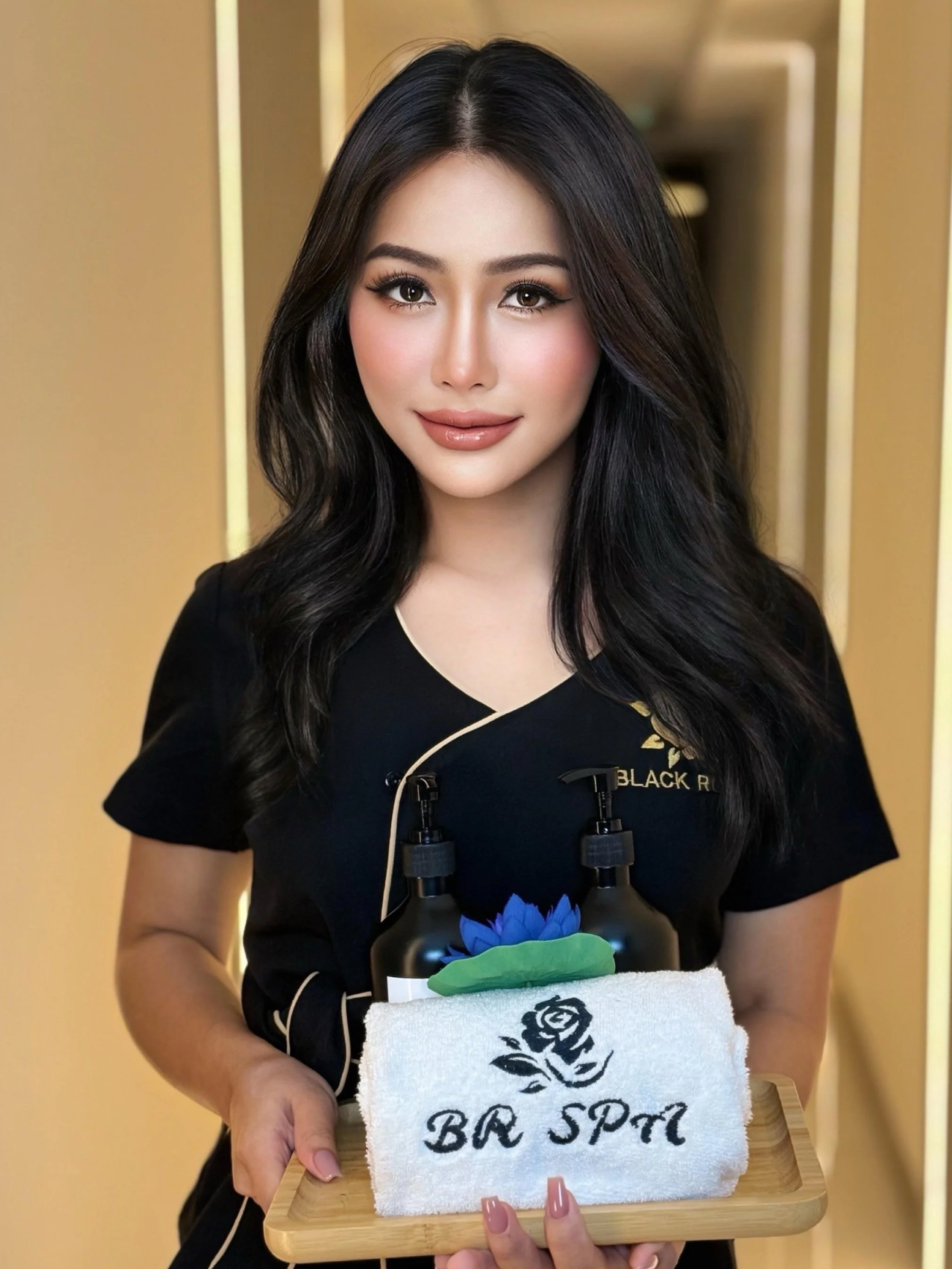 A woman with long black hair, wearing a black shirt, holding a wooden tray with a towel that has