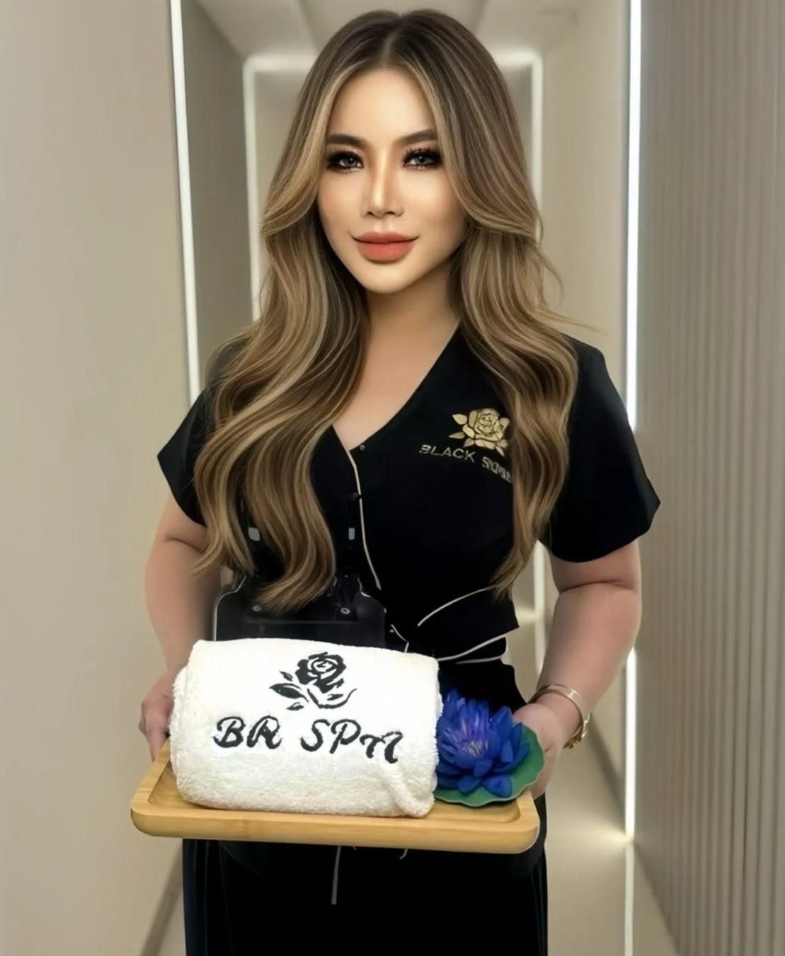 A woman with long wavy blonde hair wearing a black uniform holds a tray with a towel embroidered with a rose and the words "BA SPA" and a blue flower. She is smiling in a spa setting.