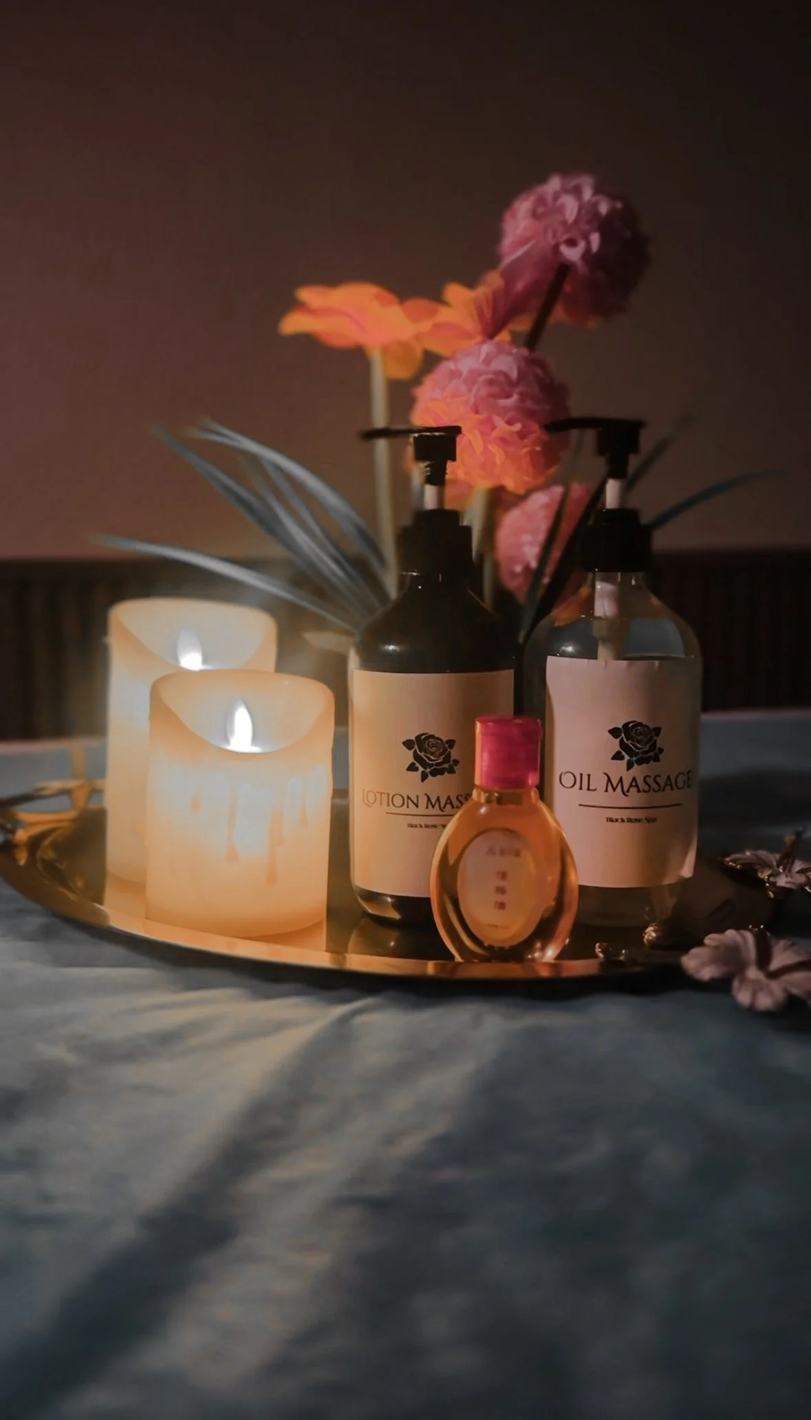 A tray with candles, lotion bottles labeled "Lotion Massage" and "Oil Massage," a small bottle, and pink flowers, all illuminated by candlelight.