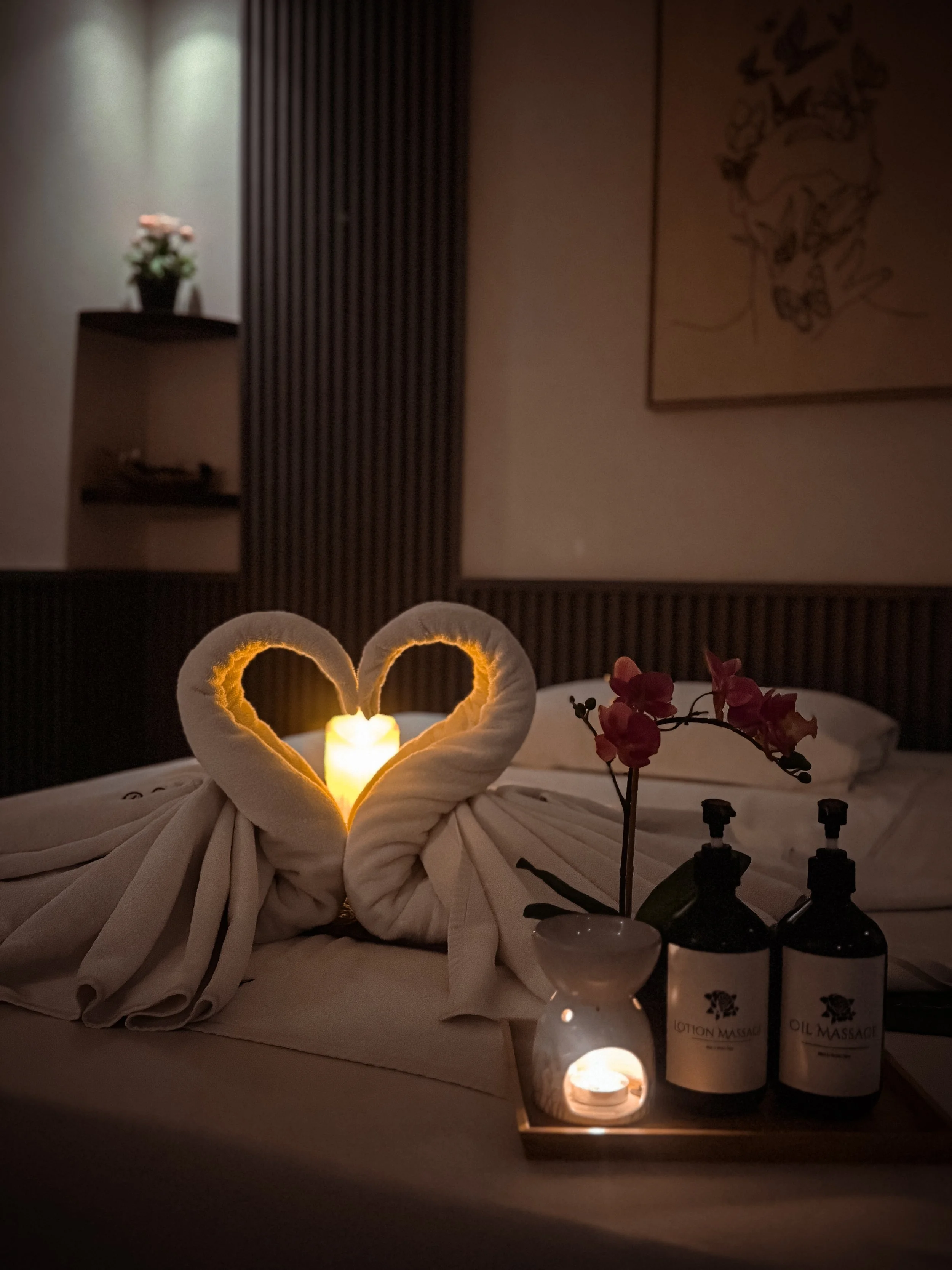 A towel folded into a swan shape with a heart in the middle, illuminated by a small candle. In the background, there is a flower arrangement on a shelf, framed art on the wall, and bottles of massage oil or lotion on a tray.