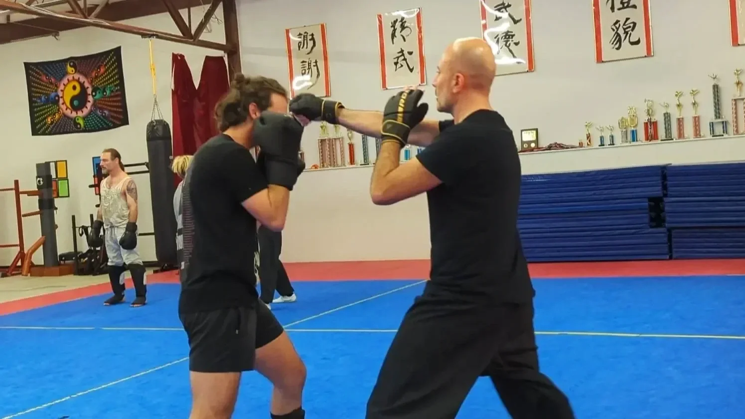 Two people practicing boxing in a martial arts gym, with a trainer demonstrating proper punching technique while a boxer blocks a punch. In the background, another person stands near training equipment.