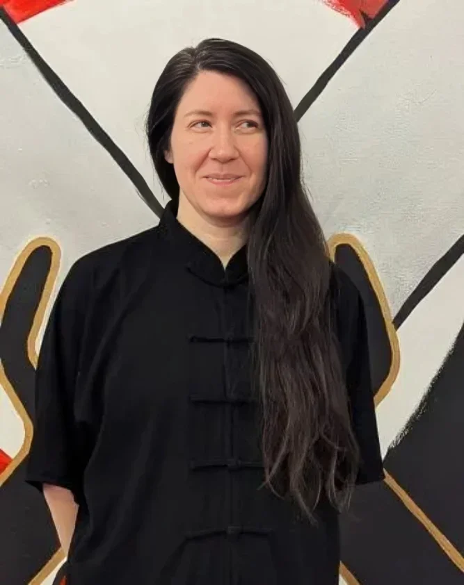 Woman with long dark hair wearing a black martial arts uniform, standing in front of a mural with black, white, red, and gold elements.