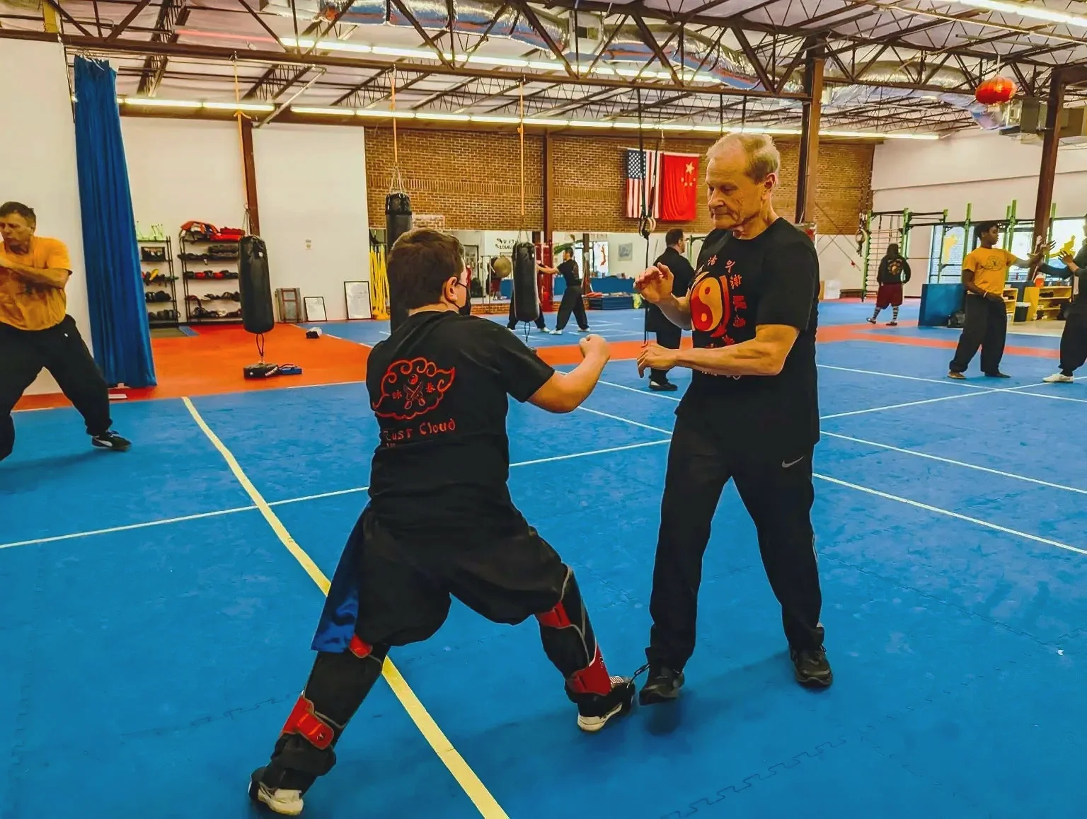 Martial arts training session in a gym with several people practicing. In the foreground, an instructor demonstrating moves to a student. The gym has blue mats, punching bags, and decorations including flags on the wall.