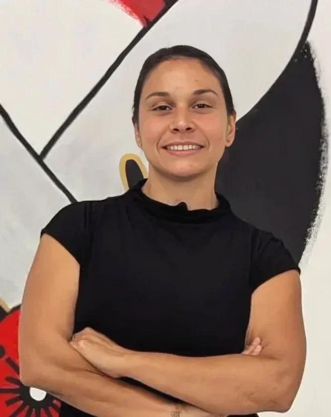 A young woman with dark hair pulled back, wearing a black t-shirt, standing with arms crossed in front of a colorful abstract wall art.