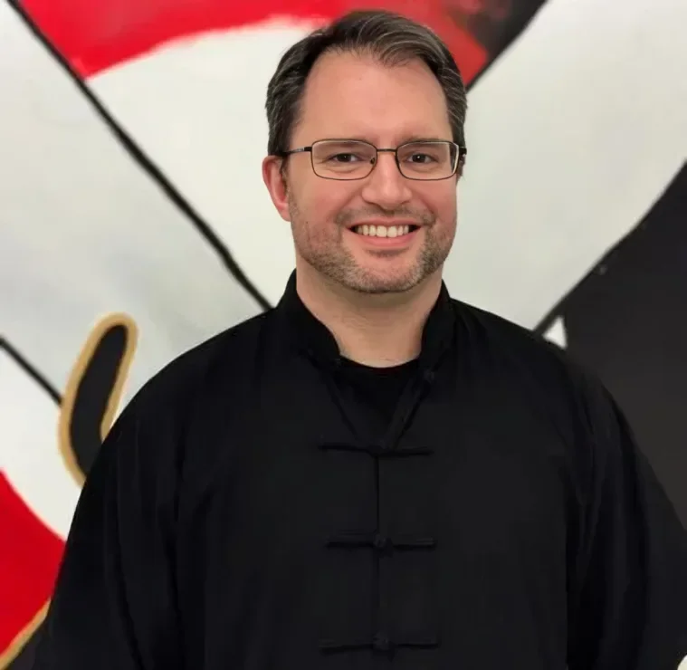 A smiling man with glasses and dark hair, wearing a black traditional Chinese martial arts uniform, standing in front of a red, white, and black background.