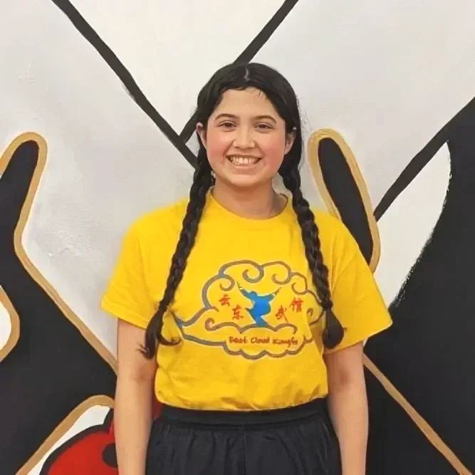 A young girl with long braided hair smiling, wearing a yellow T-shirt with a logo and text, in front of a white wall with black and gold designs.