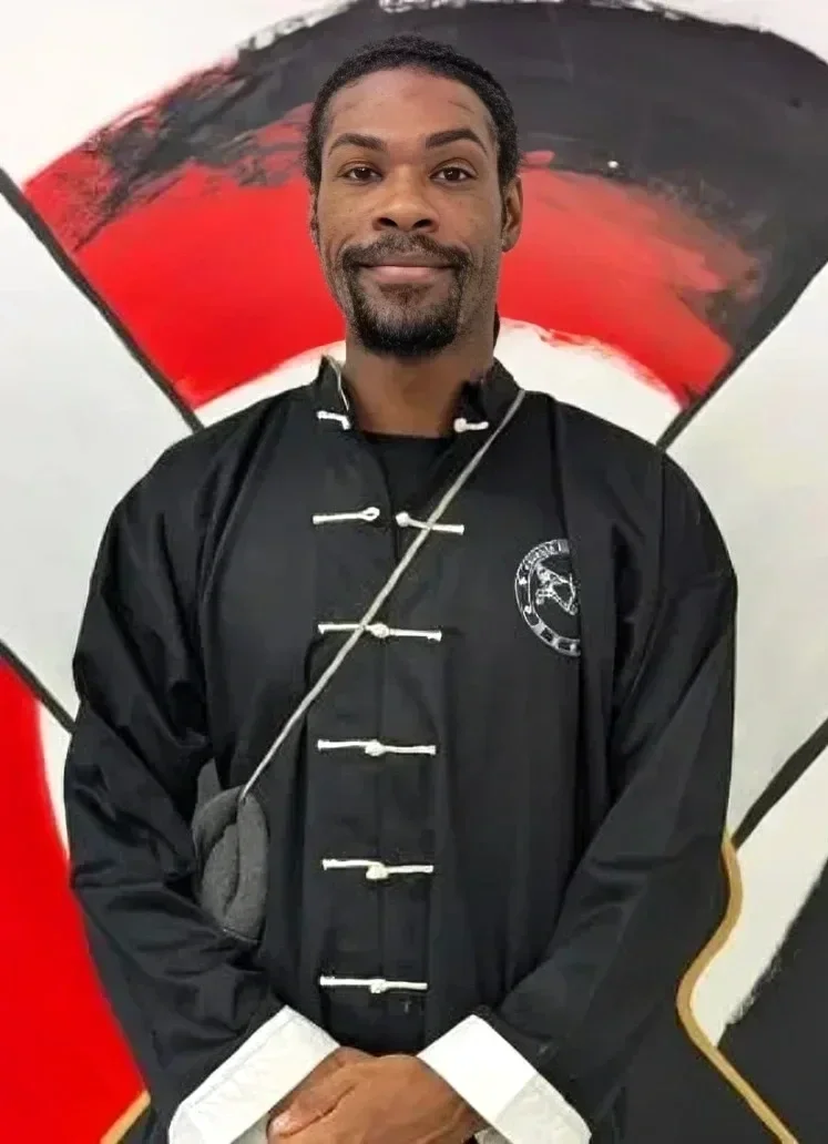 A man dressed in a black martial arts uniform stands in front of a red and black abstract background, holding his hands together.
