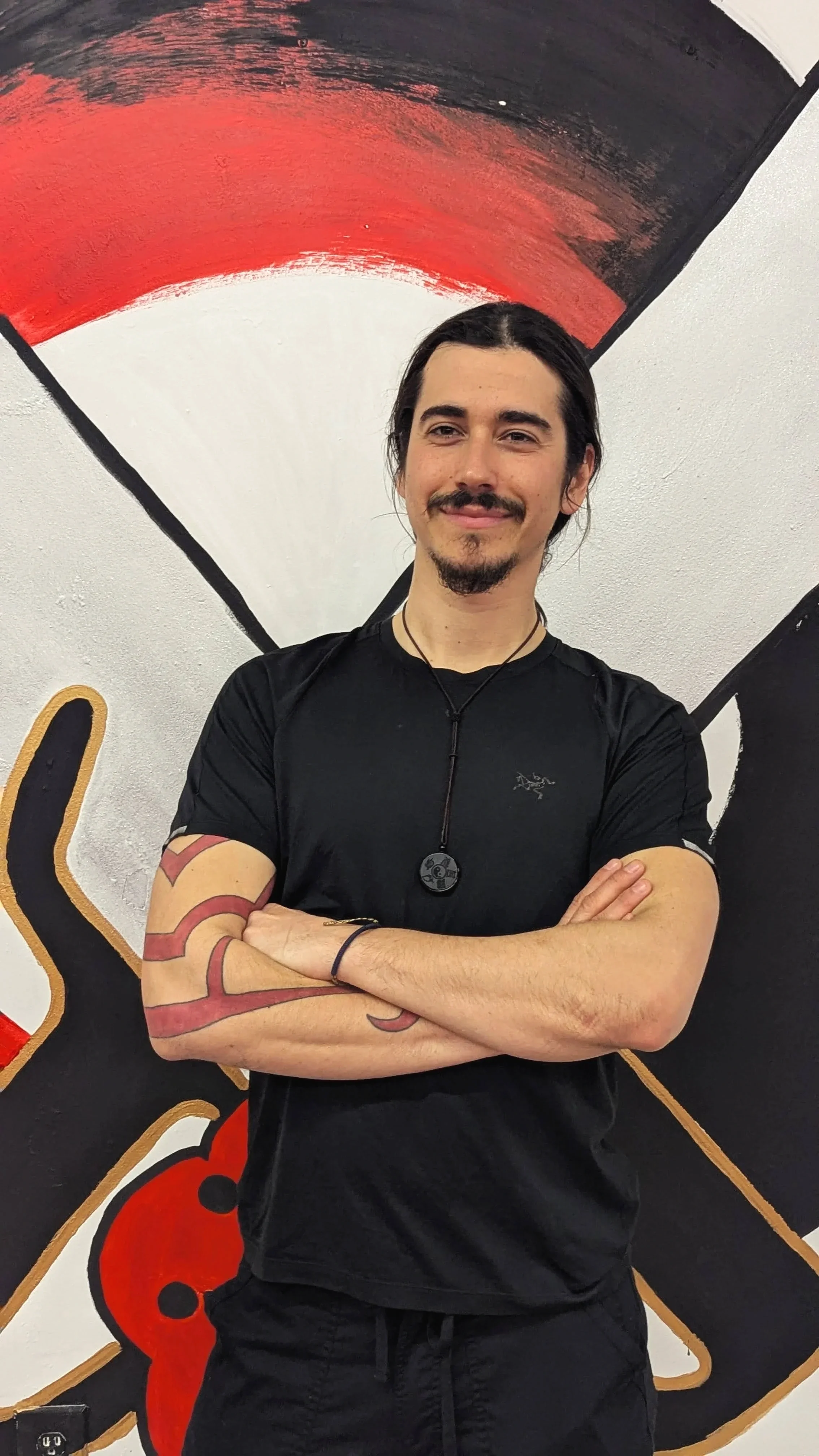 A man with long dark hair, a mustache, and goatee, wearing a black athletic shirt with a logo, and black shorts, standing with his arms crossed in front of a colorful mural.