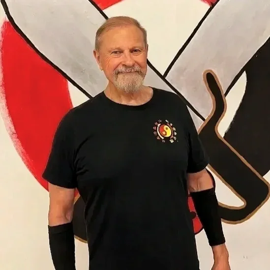 A smiling man with a beard and blonde hair stands in front of a martial arts-themed mural. He is wearing a black T-shirt with a martial arts logo and black arm sleeves.