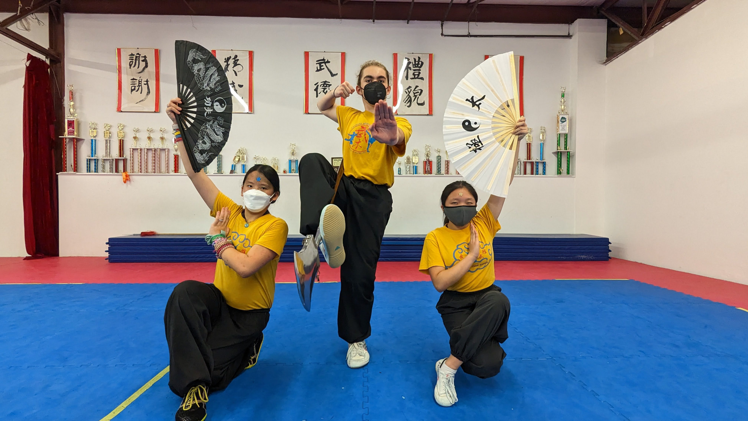 Four martial arts students posing in a dojo, wearing yellow T-shirts and black pants, holding fans with Chinese characters, making martial arts poses, with trophies and calligraphy scrolls in the background.