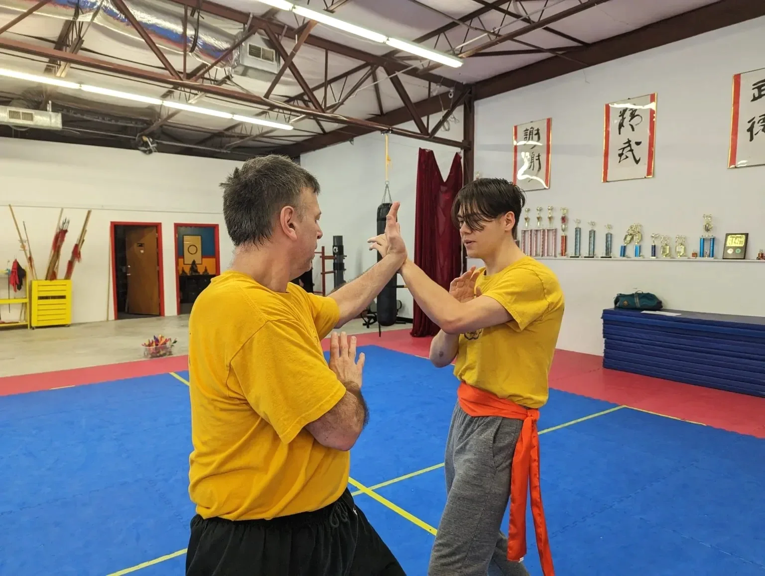 Two men practice martial arts in a dojo, wearing yellow shirts. They are facing each other, with one man's hand raised for a block while the other prepares to strike. The dojo has martial arts trophies on a shelf, calligraphy on the wall, and a punching bag in the background.