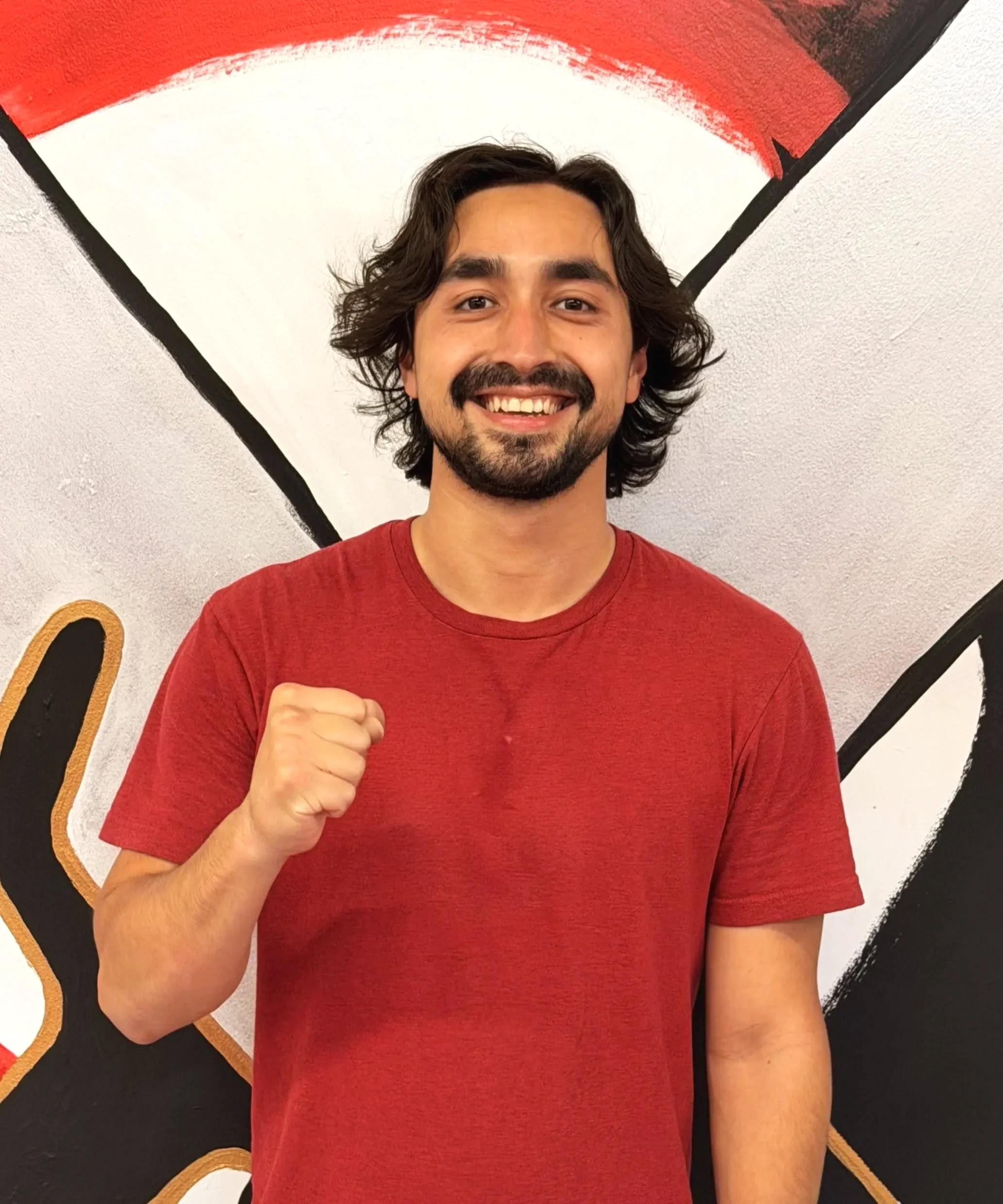 A young man with dark hair and a beard, smiling and wearing a red t-shirt, making a fist with his right hand in front of a wall with a red, white, and black design.