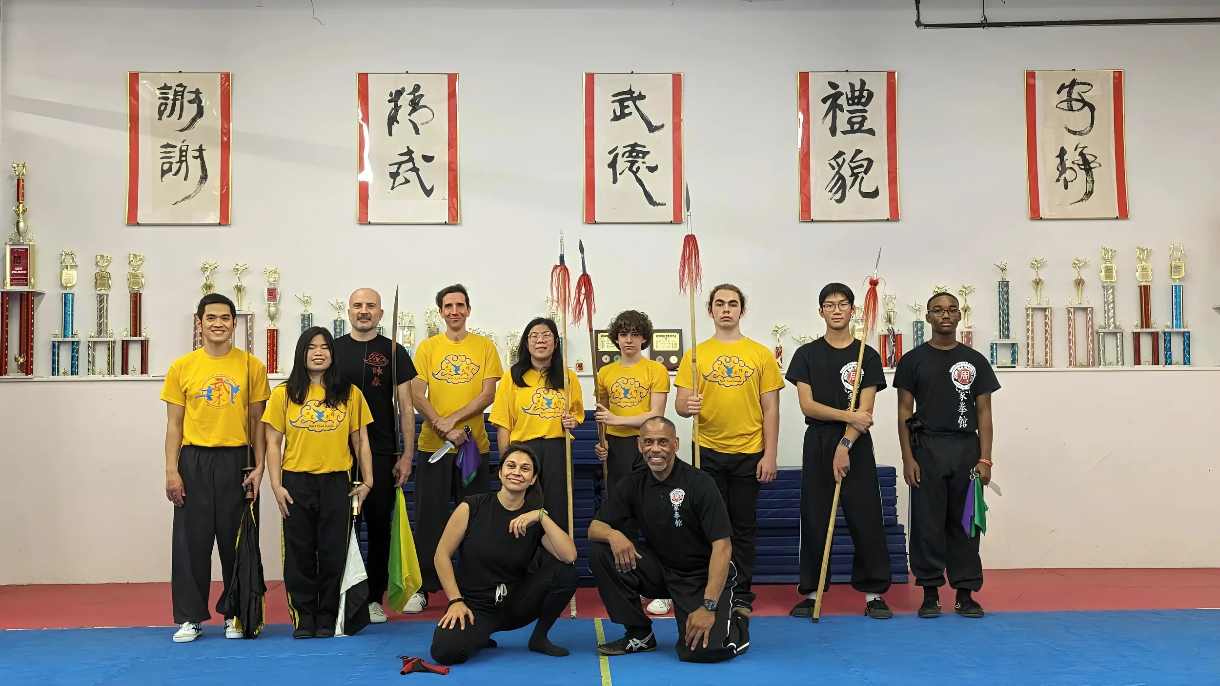 Group of martial arts students and instructors posing in a dojo with trophies and calligraphy banners on the wall.