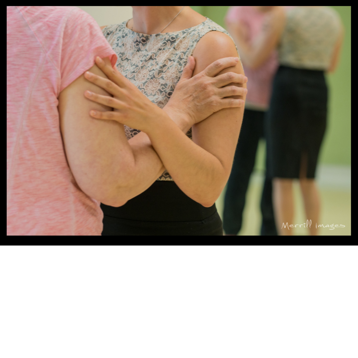 Two people holding each other's shoulders in close contact during a social gathering or dance class, with three other people blurred in the background.