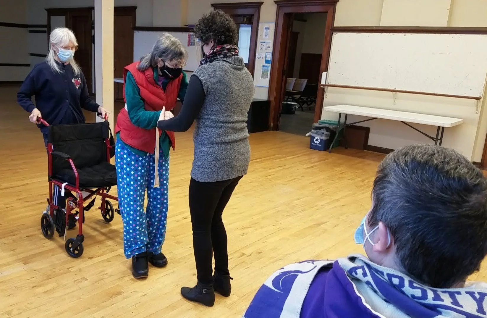 Four people in a room with wooden floors, three women and one man, all wearing masks. One woman with gray hair and glasses is using a cane and wearing colorful pajama pants with blue polka dots, holding hands with another woman who is assisting her. A woman with white hair and glasses pushes a wheelchair, and a man with brown hair and glasses is seated in the foreground wearing a mask and a sports jersey with purple and white colors.
