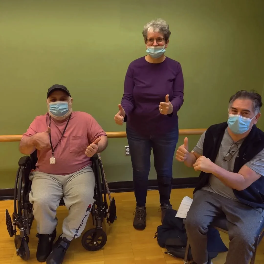 Three people wearing face masks and giving thumbs-up, two men sitting in wheelchairs and a woman standing between them.