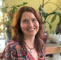 A smiling woman with shoulder-length brown hair, wearing a patterned top, standing indoors near a plant.