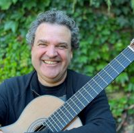 Man with curly hair smiling, holding an acoustic guitar outdoors with green foliage in the background.